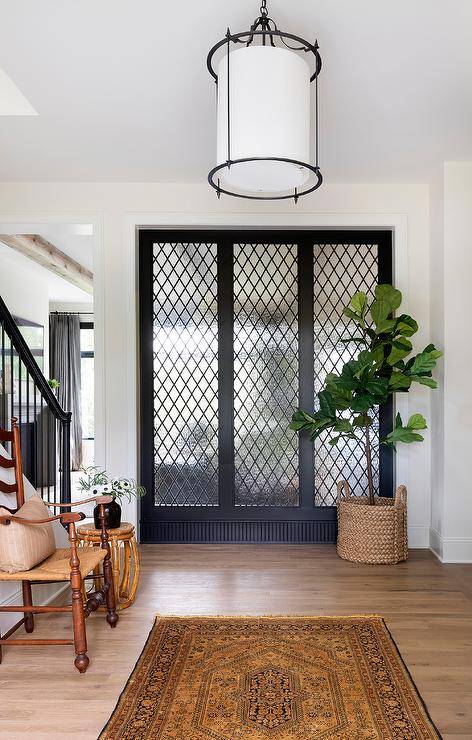 Leaded glass windows in a transitional foyer styled with a fiddle leaf fig plant in a woven basket, wood antique furniture, and a gold vintage rug. A tall ladder back wooden chair and rattan accent stool table bring an old-world feeling to the space keeping the space timeless.