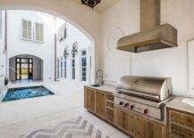 This stunning Moroccan style covered patio is equipped with a steel and brass hood fixed over an outdoor kitchen boating oak cabinets. The cabinets are finished with nickel pulls and a white stone countertop fitted with a stainless steel sink paired with a pull out faucet.