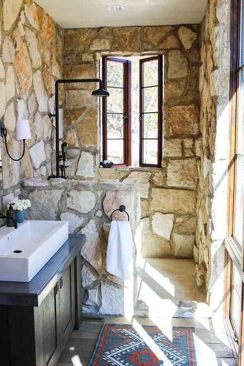 Small rustic cottage bathroom features a vanity mirror mounted to a stone covered wall beside a Reed Single Sconce and over a black washstand topped with a white overmount sink fitted with an oil rubbed bronze faucet. Rustic wood floor paneled covered in a blue and red rug leads to a walk in shower separated by a stone partition and fitted with tumbled marble brick floor tiles, a stone surround, and an oil rubbed bronze shower kit lit by natural light from a casement window.