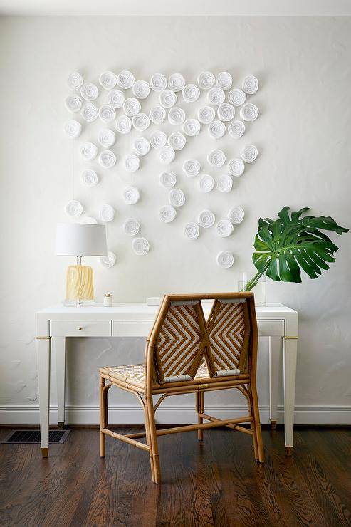 Stunning office corner designed with a white and gold desk paired with a rattan chair showcasing an amber glass lamp and a palm leaf decor. White flower accents decorate the wall above the desk for a feminine and chic finish.