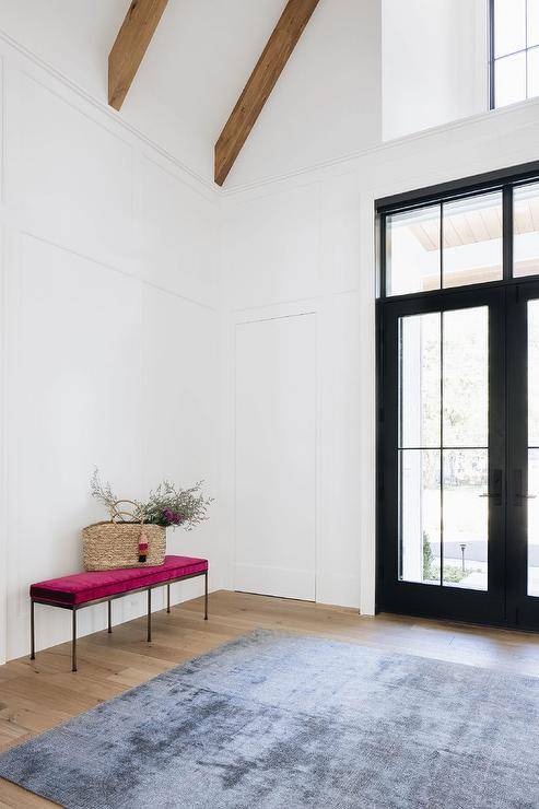 Spacious foyer features a magenta velvet bench, a gray rug, a black frame glass front door and a vaulted plank ceiling.