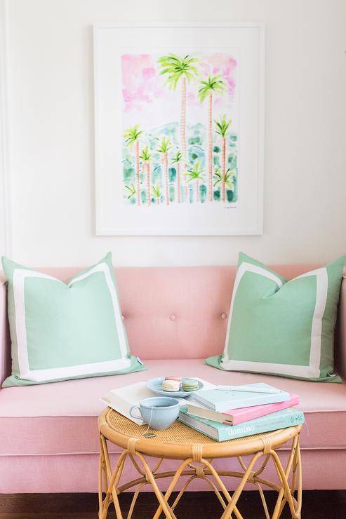 Contemporary living room furnished with a pink tufted loveseat and green pillows with white borders. The loveseat is paired with a round rattan accent table styled with pastel decor complementing a palm tree wall art.