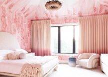 Stylish pink and cream contemporary bedroom features windows covered in pink ombre curtains hung behind a light pink velvet accent chair paired with a blue ombre stool. A cream boucle bed is placed on a cream pink rug lit by a pink feather chandelier.