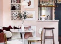 Small apartment features a white l-shaped dining bench fixed against a pink wall and topped with pink and velvet pillows. The banquette is matched with a white tulip dining table lit by a white pendant and paired with white and gold dining chairs. An inset white and black wallpapered bookshelf is fixed over the dining bench and beside stacked art.