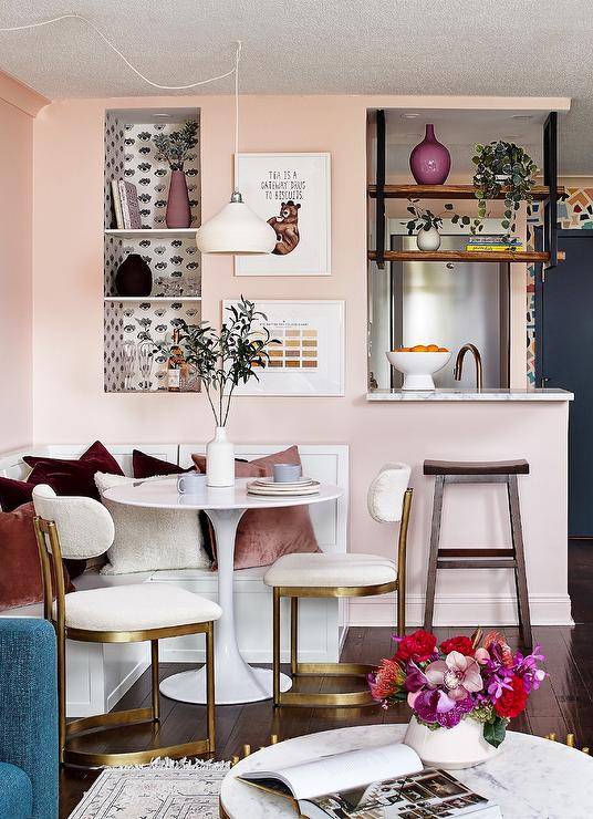 Small apartment features a white l-shaped dining bench fixed against a pink wall and topped with pink and velvet pillows. The banquette is matched with a white tulip dining table lit by a white pendant and paired with white and gold dining chairs. An inset white and black wallpapered bookshelf is fixed over the dining bench and beside stacked art.