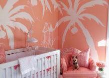 Coral pink nursery features walls painted coral, Sherwin Williams Jovial, adorned with a hand painted palm tree mural by artist EEK. A glass art deco chandelier illuminates a white crib dressed in white and pink elephant crib bedding, a Quadrille Petite Zig Zag Fabric pillow and a white and pink crib mobile next to a salmon pink chair and a Two's Company Elephant Side Table topped with pink roses as well as a gold Moroccan pouf atop a white sheepskin rug.