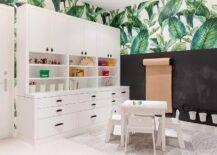 Homework room features a craft paper roll on a chalkboard wall under palm leaf wallpaper, white built in cabinets with black leather hardware and a white table with white chairs atop a beige rug.