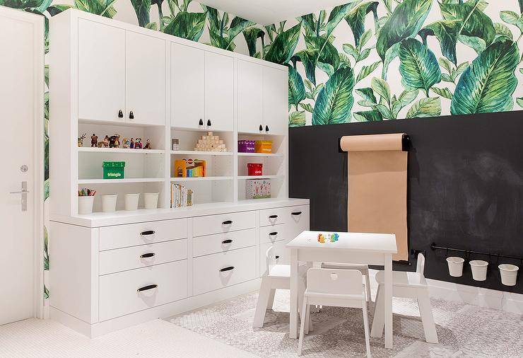 Homework room features a craft paper roll on a chalkboard wall under palm leaf wallpaper, white built in cabinets with black leather hardware and a white table with white chairs atop a beige rug.
