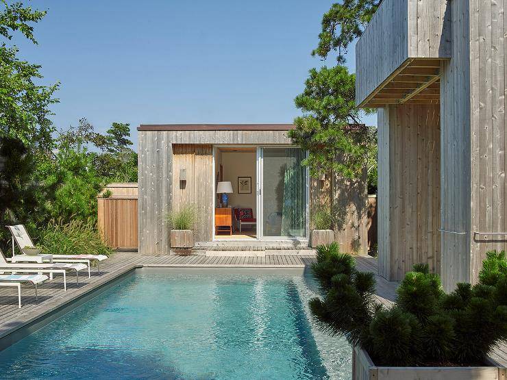 White pool loungers sit along an in ground swimming pool positioned in front of a gray plank pool house with sliding glass doors.