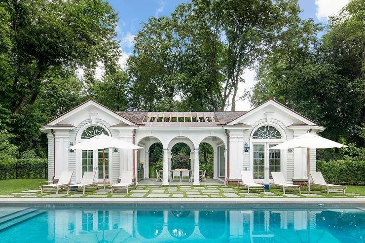 White pool loungers paired with white umbrellas face an in ground swimming pool and sit on grass lined concrete pavers leading to an expansive white pool house boasting arched doorways.