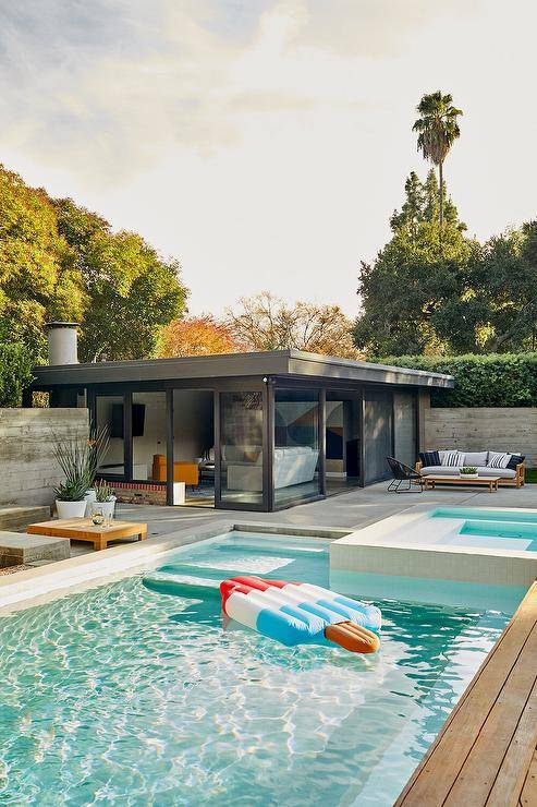 A rectangular in ground pool with a corner spa sits in front of a black pool house framed by sliding glass doors and windows.