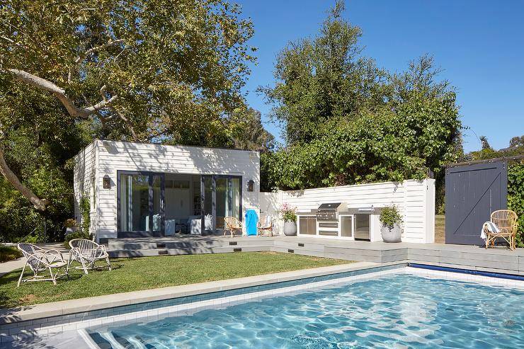 An in-ground swimming pool is fixed in front of a shiplap siding pool house accented with sliding glass doors.