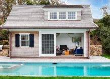 A shingled pool house is accented with black shutters and sliding glass doors.
