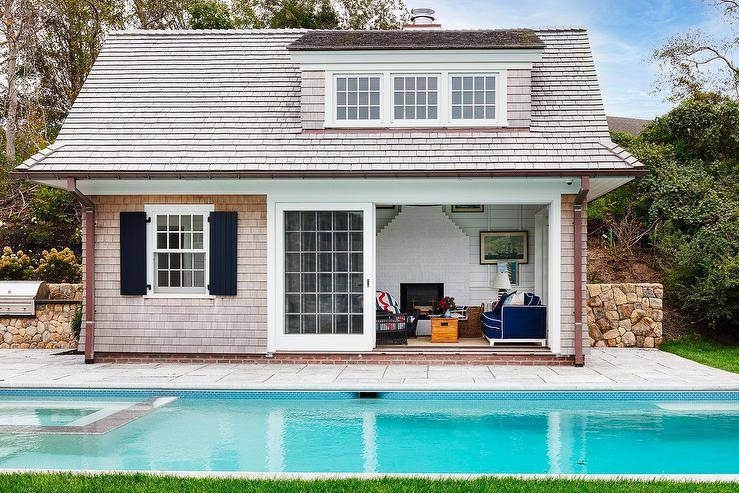 A shingled pool house is accented with black shutters and sliding glass doors.