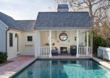 Cottage style pool house boasts a covered patio featuring a black wicker sofa and black wicker chairs placed flanking a black coffee table. A round mirror is mounted to board and batten trim over a fireplace.