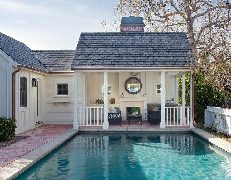 Cottage style pool house boasts a covered patio featuring a black wicker sofa and black wicker chairs placed flanking a black coffee table. A round mirror is mounted to board and batten trim over a fireplace.