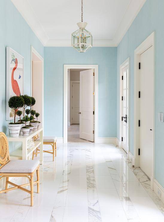 Gorgeous blue foyer features round back rattan chairs placed on either side of a white console table positioned on a white marble tiled floor beneath a flamingo art piece hung from a blue wall. The foyer is lit by a white lantern.