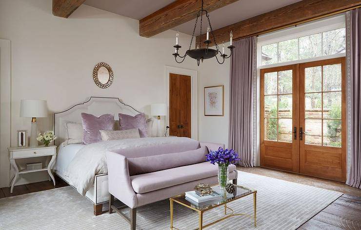 A lilac settee is placed on a light gray rug and is paired with a brass coffee table positioned at the foot of a white French bed dressed in off-white bedding complemented with lilac velvet pillows. The bed is located beneath a round gold mirror and is flanked by white French nightstands finished with alabaster lamps.
