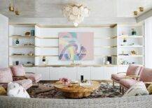 Fun spacious living room design features pink and gold chairs with a round burl wood coffee table and abstract art with white and gold built in shelves.