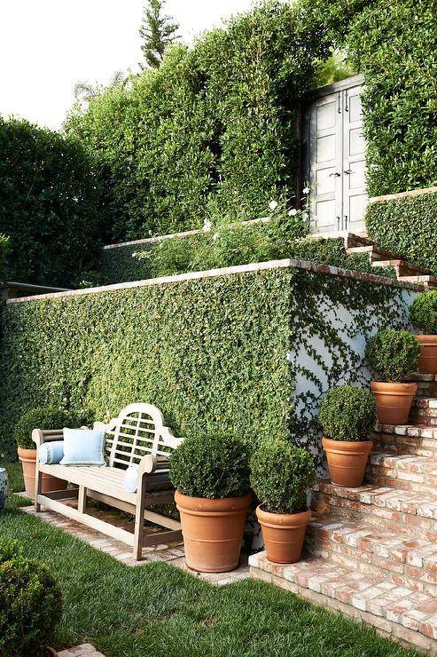 Secret Garden style backyard features a wooden bench accented with blue pillows and placed against a wall covered in moss. Brick pavers cover stairs complemented with potted plants.
