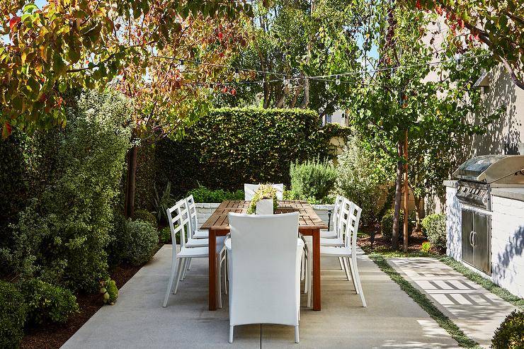 Outdoor patio design featuring tall green privacy hedges and shrubs creating an enclosure around a wood patio table and white modern chairs.