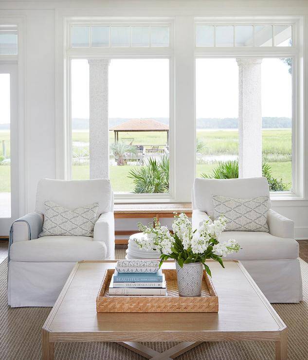 Living room features white skirted accents chairs with a tan oak coffee table that boasts a woven tray and a tan bound sisal rug, positioned in front of large bay windows to exterior