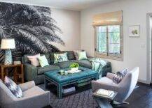 In front of a palm tree wall mural, a shimmery gray sofa with a chaise lounge is accented with a peacock blue tufted ottoman placed on a black diamond print rug and paired with gray swivel chairs.