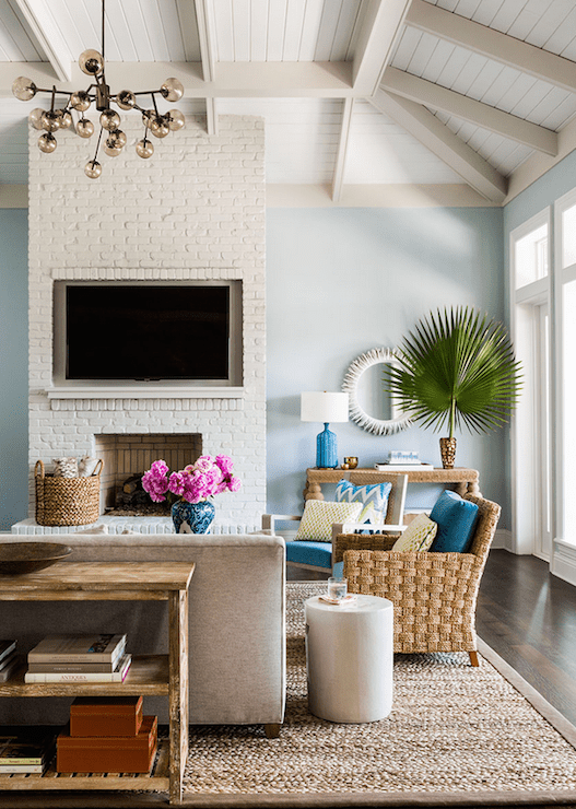 Tropical living room features paneled tray ceiling adorned with gray wood beams over a floor to ceiling white brick fireplace accented with flatscreen TV niche over white brick hearth beside a fabric wrapped console table topped with Aerin Culloden Table Lamp and brass bowls under a white sunburst mirror. Living room boasts a salvaged wood console table leaning against a velvet sofa paired with a pair of woven armchairs topped with turquoise cushions atop a bound sisal rug.