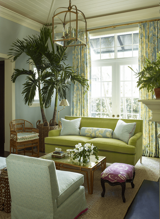 Tropical living room features an Arch Top Lantern illuminating a green velvet sofa adorned with blue pillows and cream and blue floral bolster pillows placed in front of French doors dressed in floor to ceiling cream and blue floral curtains. The green couch faces a brass and glass rectangular coffee table beside a purple ikat bench and a blue slipper chair upholstered in China Seas Java Java Fabric atop a diamond seagrass rug.