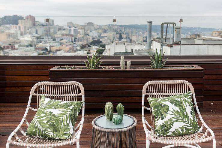 Two rope outdoor chairs with palm leaf pillows on an outdoor dark stained oak patio with a city view. A reeded outdoor accent table is styled between the two chairs displaying succulent decor.