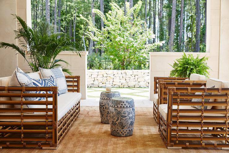 Brown teak and rattan outdoor sofas with blue pillows face a set of blue and white ceramic stools atop a tan rug.