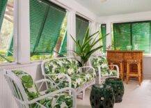 Fresh air streams into a sunroom through green shutters open above a white rattan sofa and white rattan chairs accented with white and green floral cushions and placed in front of beadboard trim. The chairs are matched with green rope stools, while wicker backless stools sit at a tiki bar.