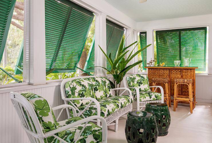 Fresh air streams into a sunroom through green shutters open above a white rattan sofa and white rattan chairs accented with white and green floral cushions and placed in front of beadboard trim. The chairs are matched with green rope stools, while wicker backless stools sit at a tiki bar.