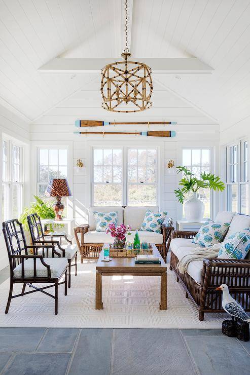 A white rug covers slate mudroom tiles and sits beneath brown rattan sofas accented with white cushions and white and blue pillows. The sofas are positioned facing a gold chinoiserie coffee table matched with two brown chairs lit by rattan light pendant hung from a vaulted plank ceiling. Decorative oars are mounted to shiplap trim over windows.