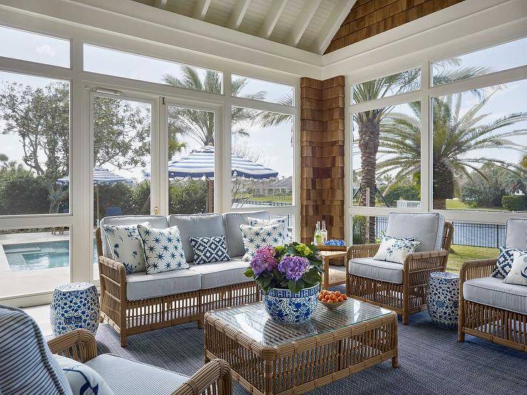A light brown rattan sofa accented with blue pinstripe cushions sits in a sunroom on a blue chevron print rug facing a rattan coffee table and paired with a blue chinoiserie drum stool and side-by-side brown rattan chairs.