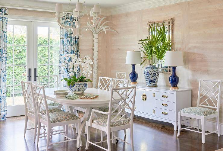 Cottage dining room furnished and styled with a white oval dining table and white lattice chairs with animal print cushions. A white palm tree floor lamp is styled in the corner against a wall with tan walls. A white chinoiserie credenza features dark blue lamps and a bamboo mirror adding an eye-catching appeal to the dining space design.