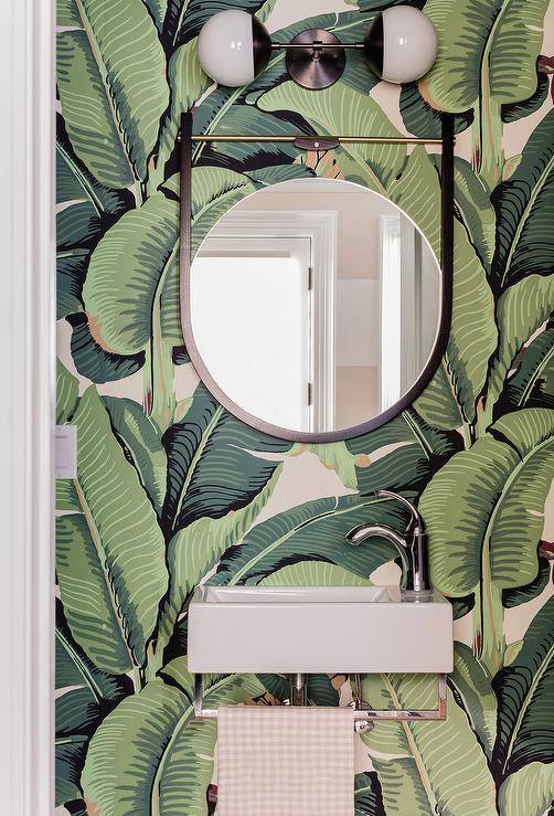Jungle palm leaf wallpaper in a powder room transforms a look into a lush, tropical escape. Wall mount sink with a mod chrome faucet complements a chrome towel rack on the base of the sink under a round black mirror lit by a two-light linear globe sconce.