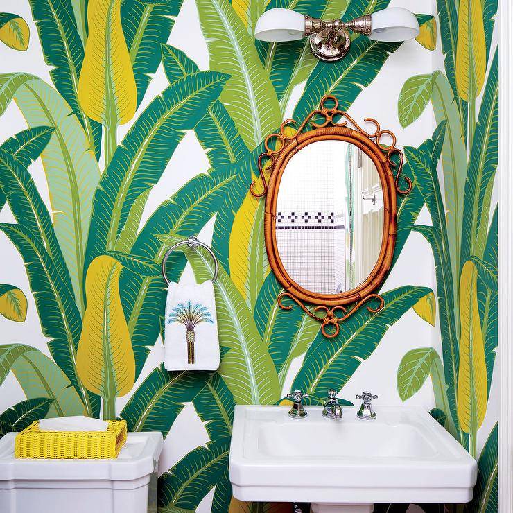 Schumacher Tropical Isle Wallpaper covers the wall of this gorgeous yellow and green powder room boasting an oval rattan mirror mounted beside a polished nickel towel ring over a pedestal sink lit by a 2-light nickel sconce.