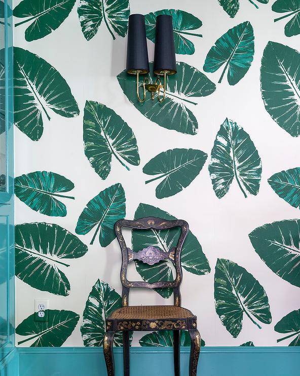 A vintage black chair sits under black sconces on green tropical print wallpaper with turquoise blue moldings.