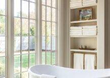 A row of tall windows are positioned behind a vintage white cast iron bathtub placed in front of built-in shelves.