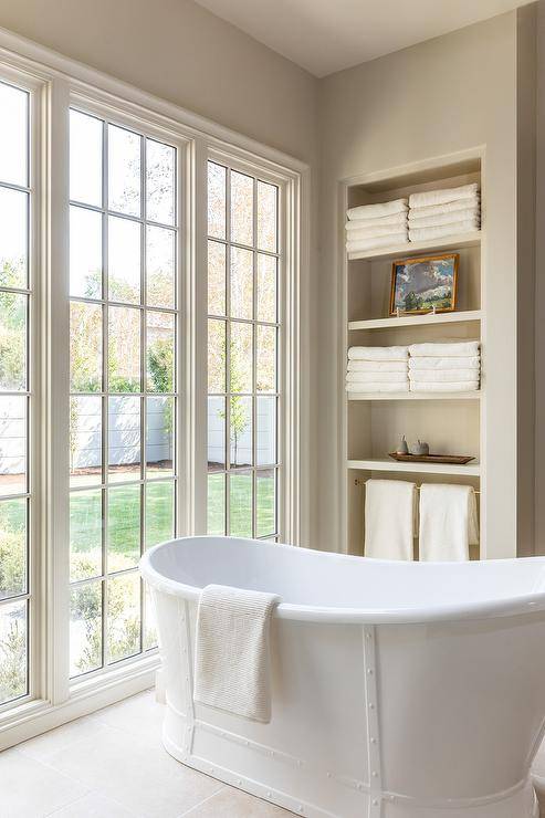 A row of tall windows are positioned behind a vintage white cast iron bathtub placed in front of built-in shelves.