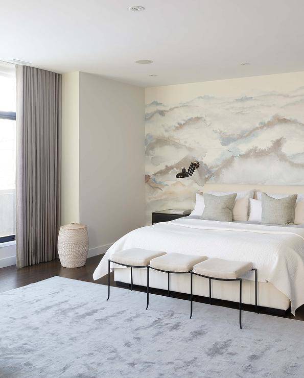 Elegant bedroom showcases a stunning watercolor wall mural located behind a cream upholstered bed topped with cream and gray bedding. A black accordion sconce is mounted above a black nightstand, while a triple seat black and gray bench is placed on a gray rug at the foot of the bed,