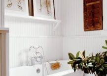 White cottage bathroom features freestanding vintage tub, polished nickel floor-mount tub filler, white beadboard walls with chair rail and white shelf filled with art over tub.