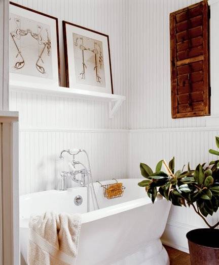 White cottage bathroom features freestanding vintage tub, polished nickel floor-mount tub filler, white beadboard walls with chair rail and white shelf filled with art over tub.