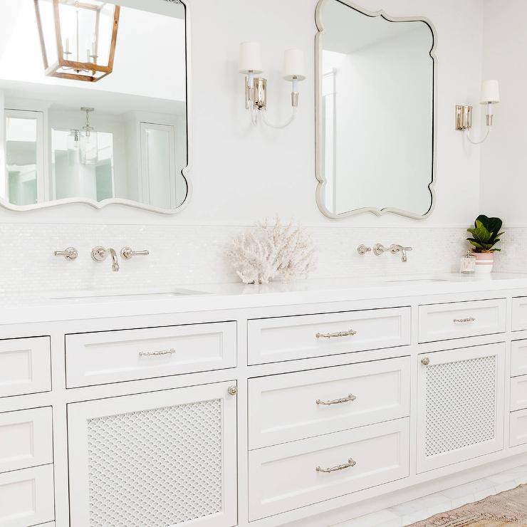 Luxurious white seaside bathroom boasts white French mirrors flanked by glass and nickel sconces and mounted over white glass mosaic backsplash tiles holding polished nickel wall mount faucets above a white dual bath vanity finished with polished nickel pulls.