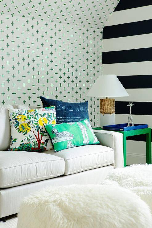 Green and blue pillows sit atop a white couch placed in front of a wall covered in green plus print wallpaper, as a wicker lamp and a blue tray sits on a green parsons end table placed beside a white and black striped accent wall.