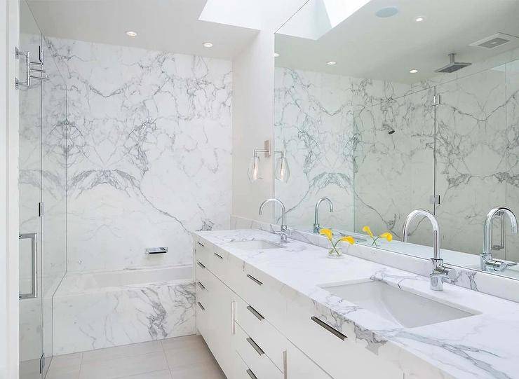 A marble clad drop-in bathtub with a wall mount tub filler is fixed against a marble slab backsplash. A skylight is located over a full-length frameless vanity mirror hung above a white dual washstand finished with flat front cabinets and a marble countertop with polished nickel gooseneck faucets.