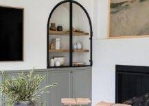 An inset arched cabinet with glass and metal doors is fixed over gray built in TV cabinets.