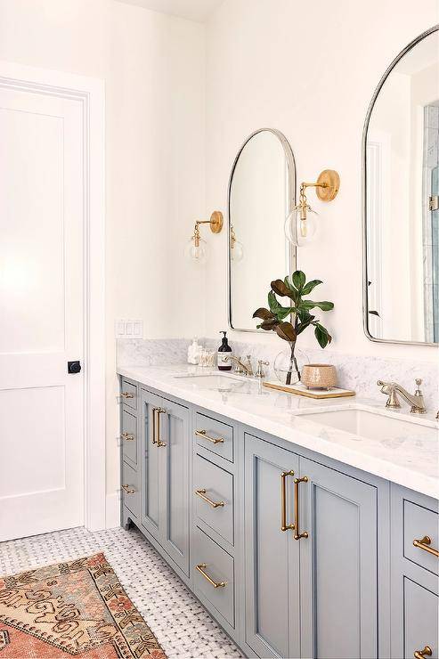 Bathroom features a double gray bath vanity with brass pulls, nickel mirrors flanked by glass and brass sconces, a marble countertop with nickel faucet, and a vintage rug on marble basketweave floor tiles.