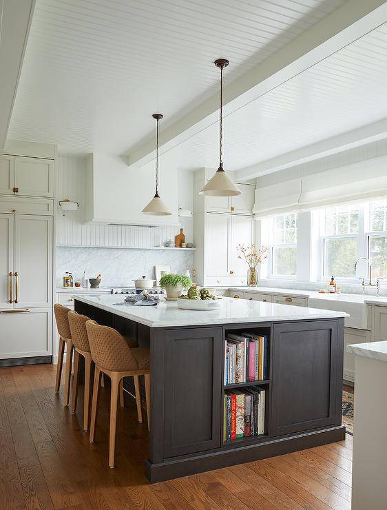 Stacked cookbook shelves are fitted to a black kitchen island topped with a marble countertop and matched with brown woven stools lit by white and black vintage lanterns hung from a beadboard ceiling.